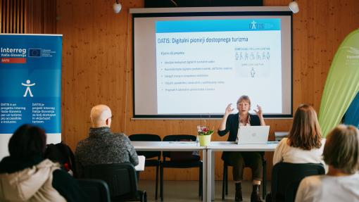 A female lecturer sitting behind the table talking to the audience. Projection screen and roll-up of DATIS project next to her.