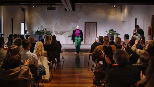 A model confidently walks down a runway in a stylish outfit, wearing green trousers, a magenta top, and a black jacket, as an engaged audience looks on and applauds. The setting is an intimate indoor venue with a rustic backdrop, and several attendees capture the moment on their phones, highlighting the event's vibrant and inclusive atmosphere.
