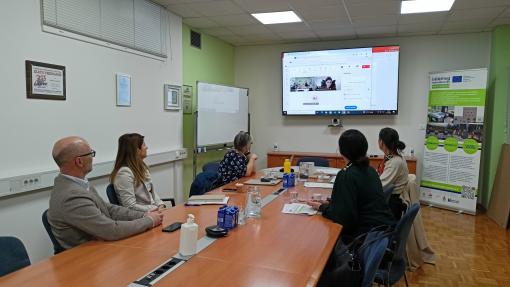 Skupina ljudi sedi za konferenčno mizo v sejni sobi, kjer spremljajo virtualno srečanje na zaslonu. Na mizi so steklenice vode, dokumenti in digitalne naprave, kar nakazuje na strokovno razpravo in sodelovanje pri projektu.