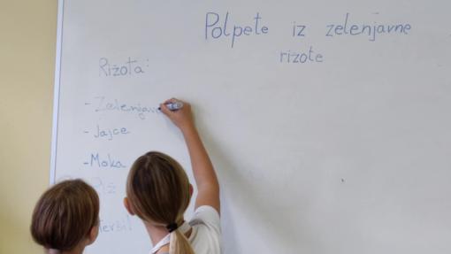 Two young girls standing in front of a whiteboard, writing down ingredients for "Vegetable Risotto Patties" during a home economics class. They are wearing aprons and working together to list the recipe.
