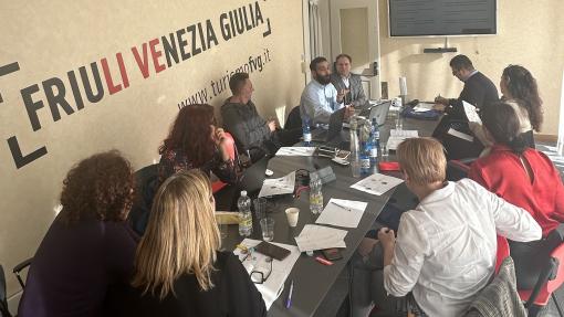 A group of people in a work meeting sitting behind a table infront of a screen. The sign on the wall behind says:"Friuli Venezia Giulia"