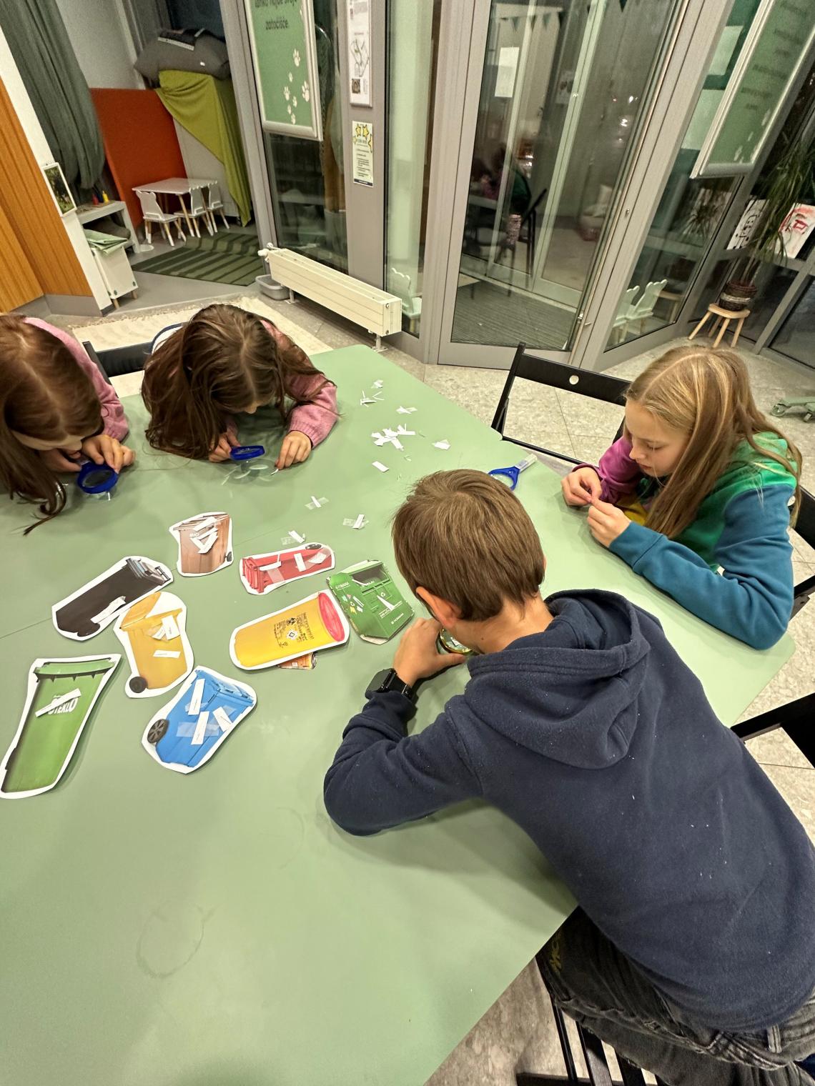 A group of children sitting around a table engaged in an activity about recycling. They are sorting and matching waste items with corresponding recycling bins, focusing intently on the task in a family activity center.