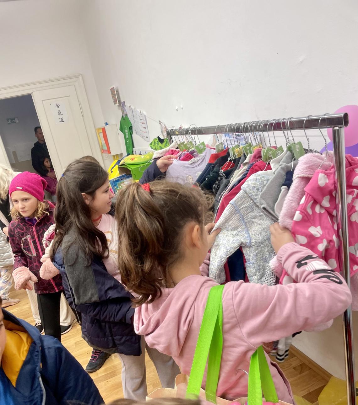 A group of children browses through a rack of colorful clothes in a bright, indoor space. The children are engaged in selecting garments.