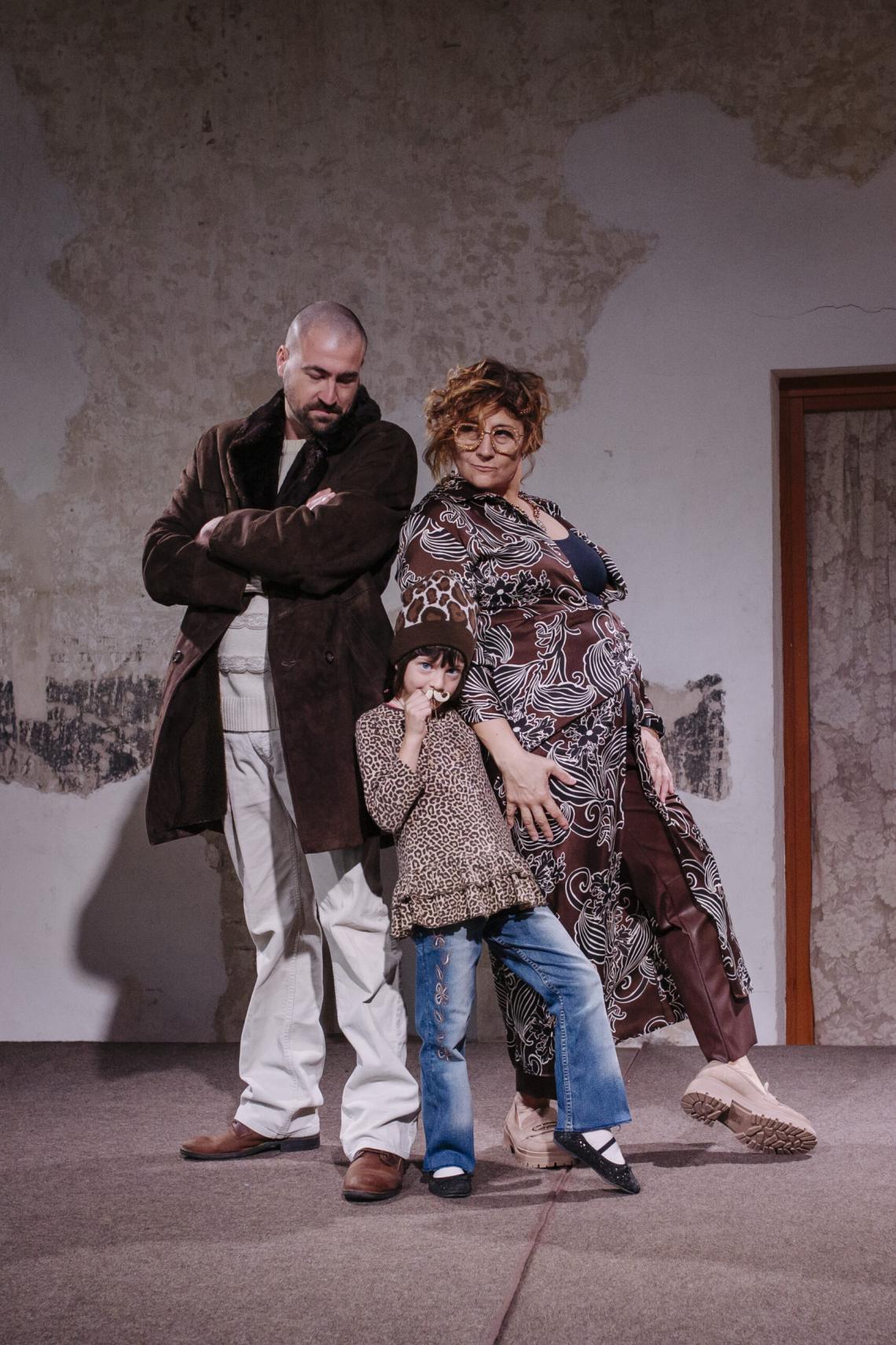 A stylish trio poses confidently on a runway in a rustic setting. The adult man, dressed in a brown coat and neutral tones, stands with arms crossed. The woman beside him wears a bold, patterned outfit and round glasses, exuding confidence. A young child stands in front of them, wearing a leopard-print top and jeans, playfully adding charm to the group.