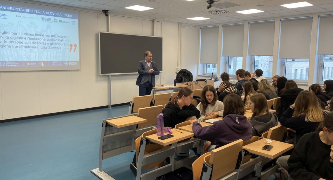 A professor standing in the class room, lecturing to students. Some are turned to each other and discusing or doing common exercises for the lecture.