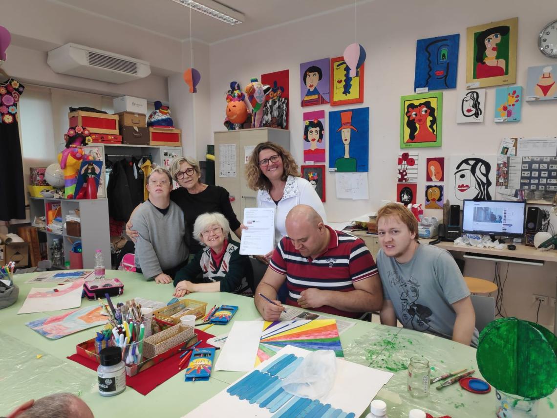 Members of Coop Mosaico sitting at a table in their creative room. A man is fillin out a paper form. Three people are standing next to him, smiling.
