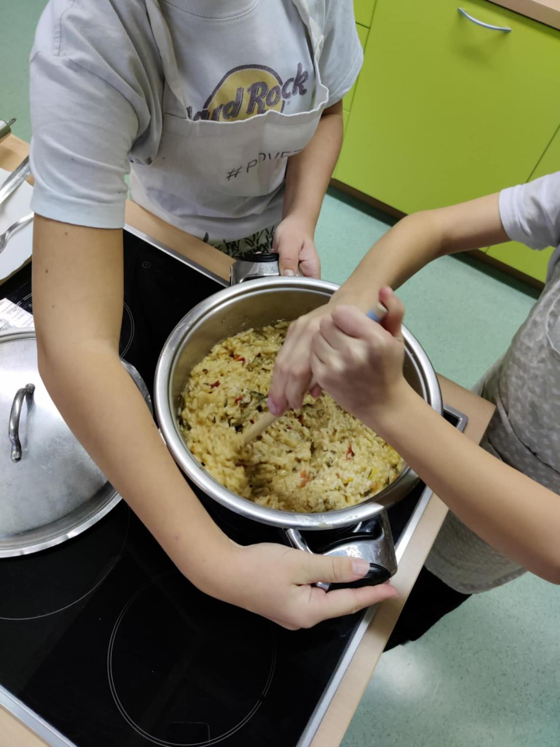 Close-up of two students stirring a pot of vegetable risotto on a stovetop during a cooking class.